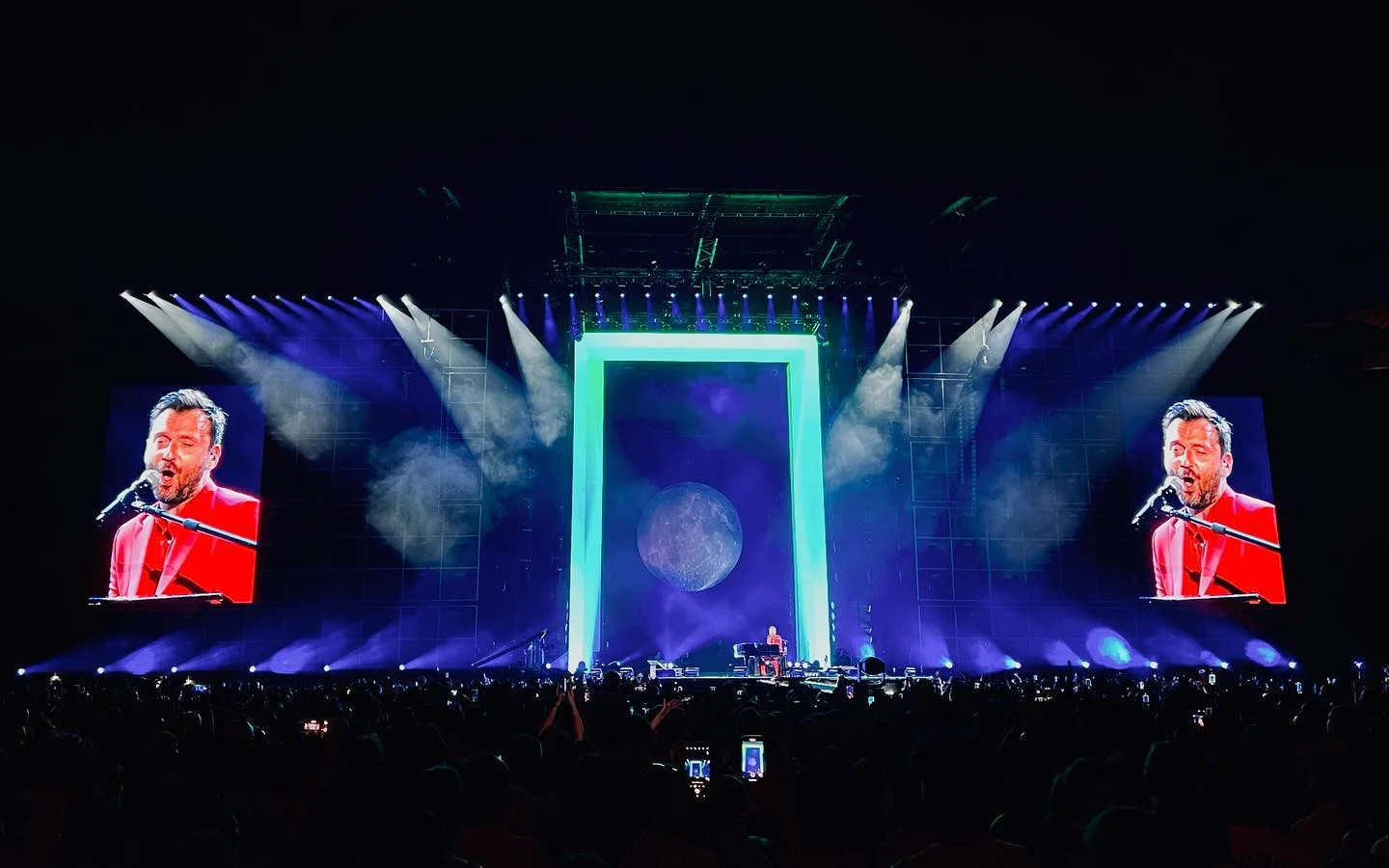 Il portale illuminato di verde attorno a una proiezione della luna, Cesare Cremonini al pianoforte sugli schermi laterali – Cesare Cremonini Stadi 2022, Italia – scenografia per concerti