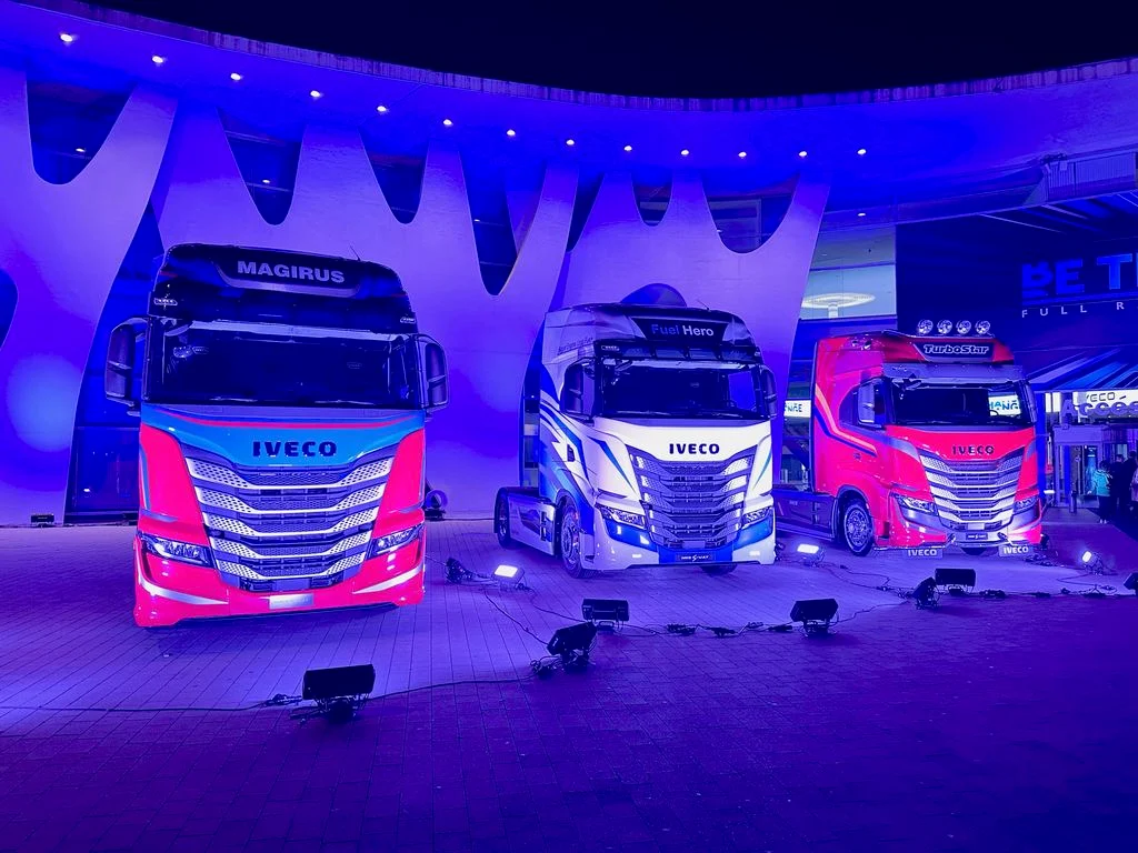 Tre cabine di camion con livree rosa, bianca e blu sotto una copertura ondulata illuminata di viola – Lancio IVECO Be The Change, Barcellona, Spagna, 2023 – progettazione di eventi