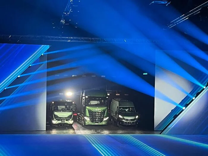 Tre veicoli Iveco verdi che escono da un'apertura nella parete LED sotto fasci di luce blu – Lancio IVECO Be The Change, Barcellona, Spagna, 2023 – progettazione di eventi