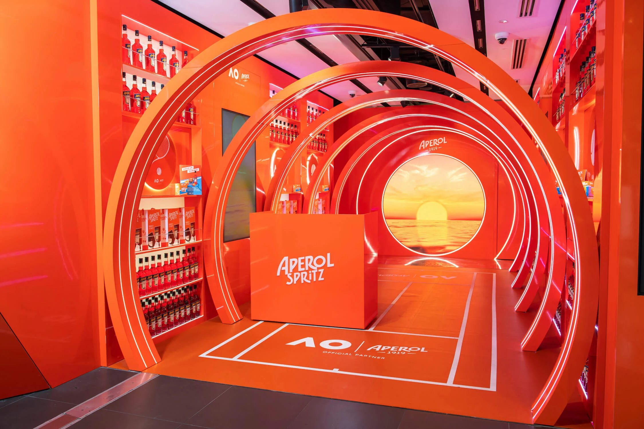 Vista angolata del tunnel di archi arancioni vuoto con il bancone Aperol Spritz e le linee del campo – Aperol Pop-Up all'aeroporto di Melbourne, Australia, 2024 – retail design