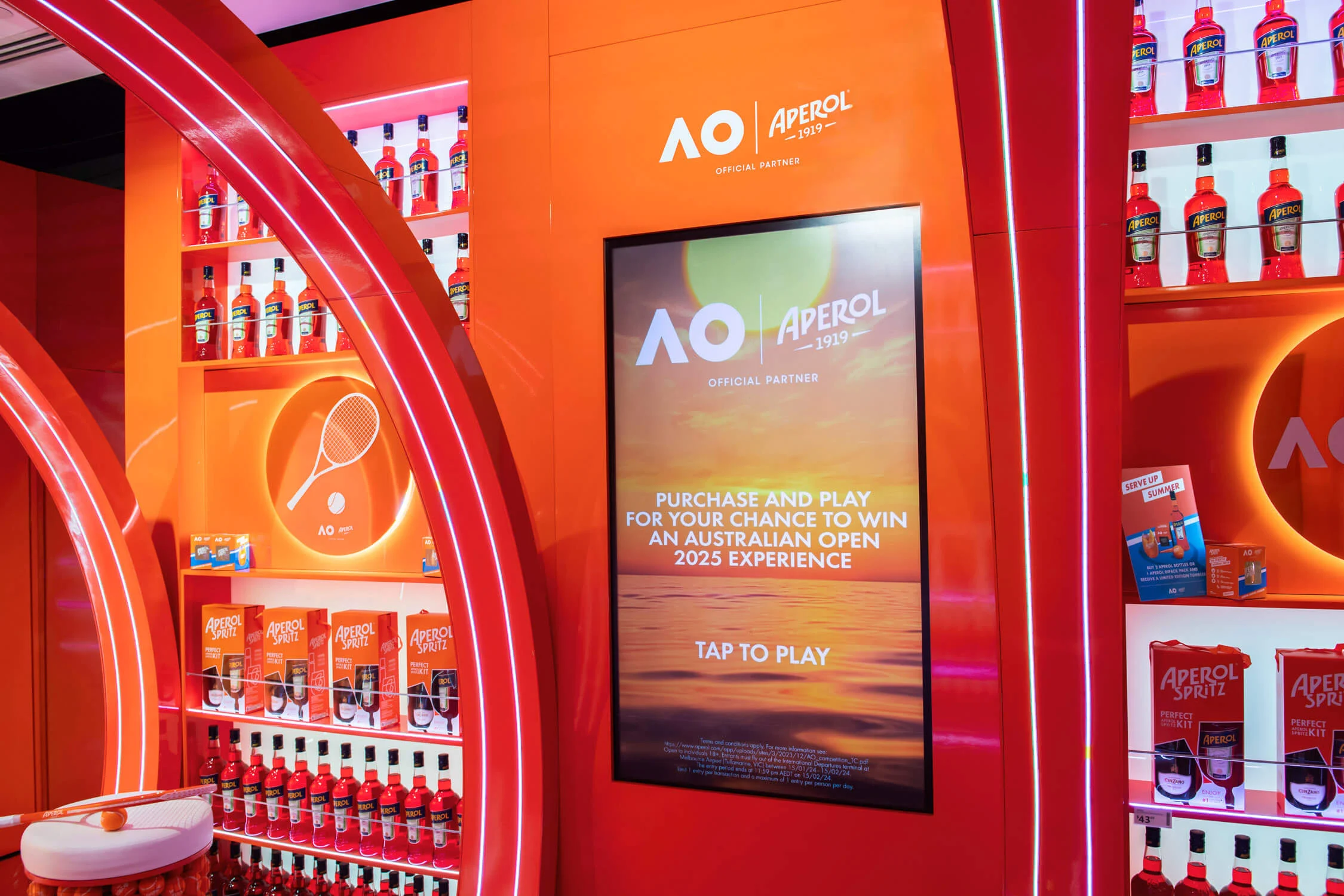 Touchscreen che invita i viaggiatori a giocare per vincere un'esperienza all'Australian Open, tra scaffali arancioni di bottiglie – Aperol Pop-Up all'aeroporto di Melbourne, Australia, 2024 – retail design