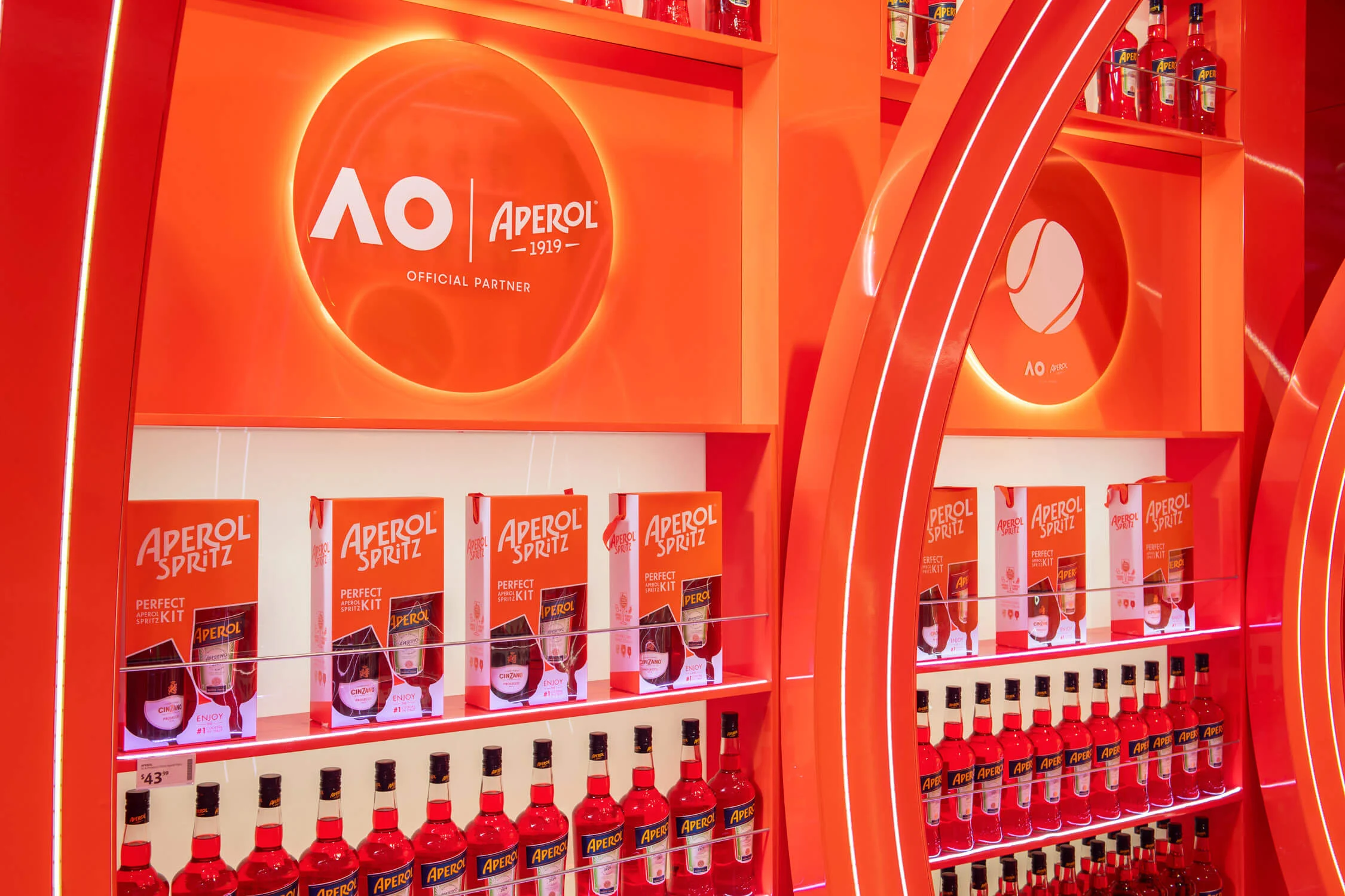 Dettaglio di scaffali con bottiglie e confezioni regalo Aperol sotto il logo retroilluminato Australian Open e Aperol – Aperol Pop-Up all'aeroporto di Melbourne, Australia, 2024 – retail design