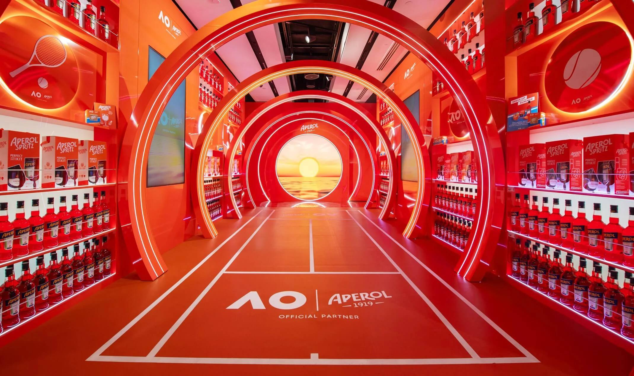 Shop-in-shop Aperol arancione: un tunnel di archi illuminati su un pavimento da campo da tennis che porta a uno schermo con un tramonto – Aperol Pop-Up all'aeroporto di Melbourne, Australia, 2024 – retail design