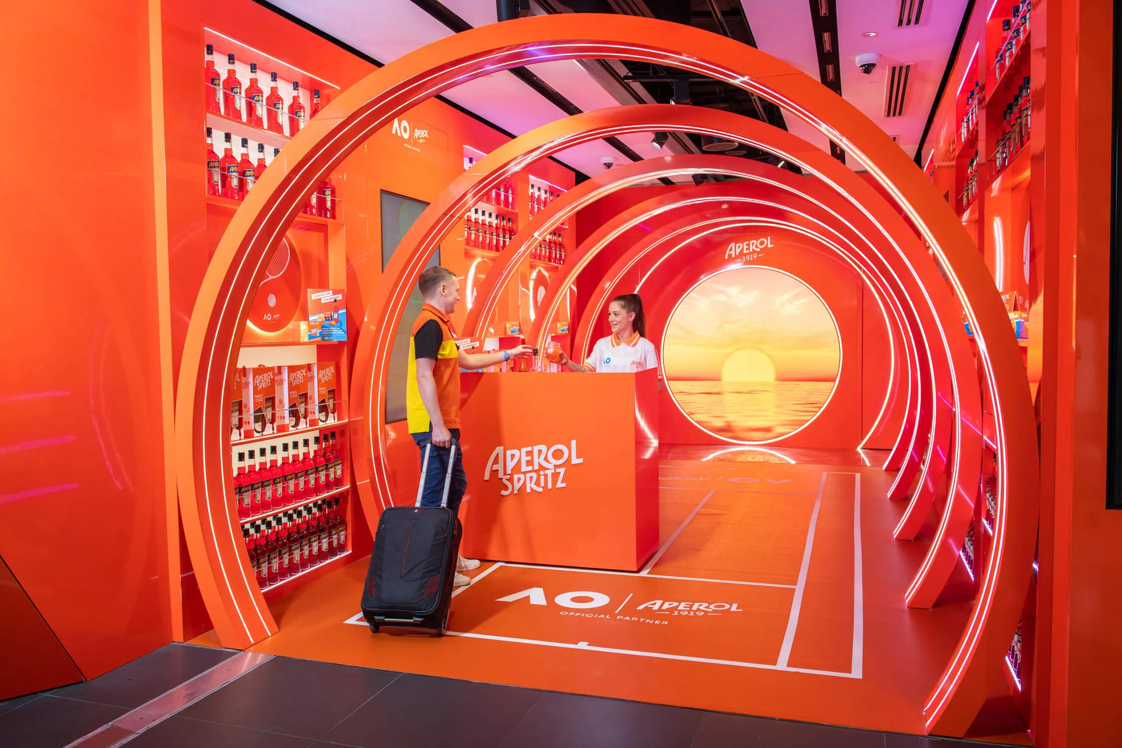 Viaggiatore con valigia al bancone Aperol Spritz dentro il tunnel di archi arancioni – Aperol Pop-Up all'aeroporto di Melbourne, Australia, 2024 – retail design