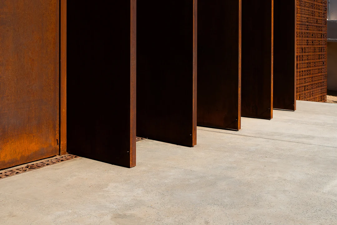 Dettaglio di lamelle metalliche color ruggine che proiettano ombre nette su un pavimento in cemento – Design Space AlUla, Arabia Saudita, 2024 – architettura