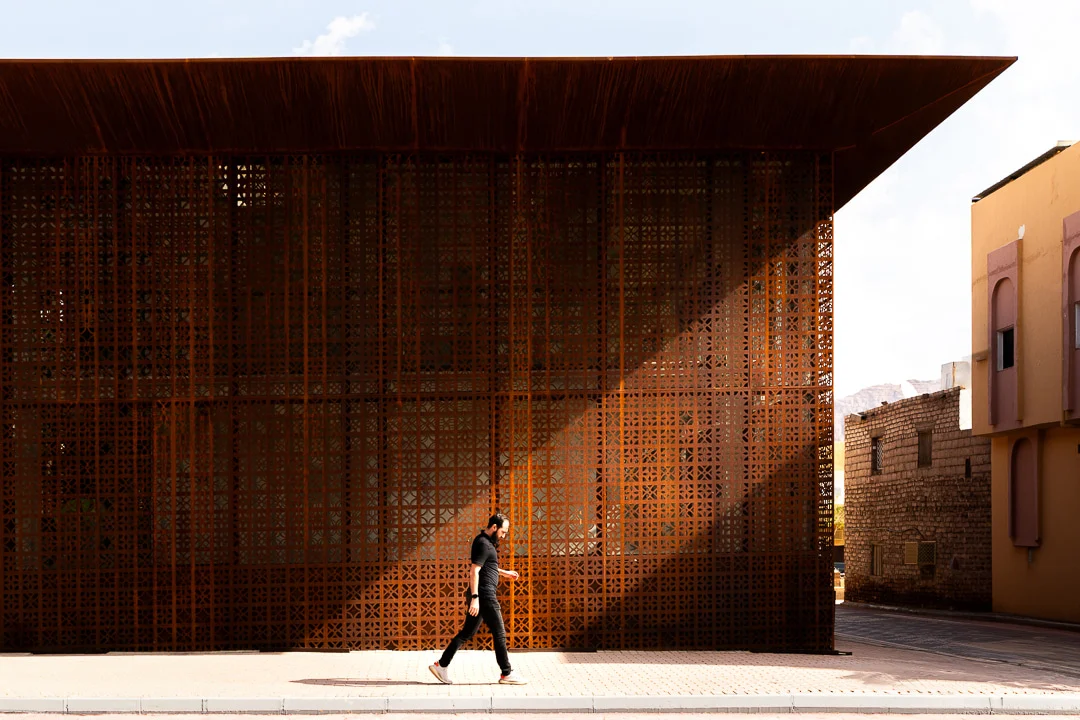 Persona che cammina lungo la facciata a schermo forato nella luce radente, sotto il profondo sbalzo del tetto – Design Space AlUla, Arabia Saudita, 2024 – architettura