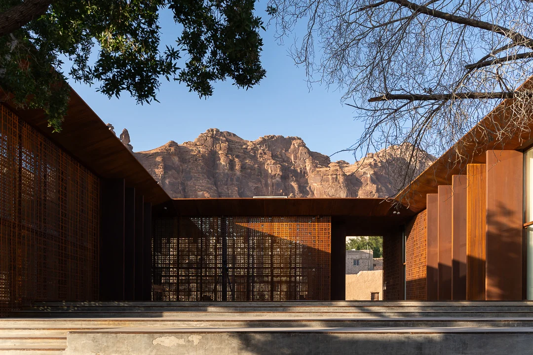 Cortile racchiuso da schermi e lamelle in metallo color ruggine, con le falesie rocciose di AlUla dietro – Design Space AlUla, Arabia Saudita, 2024 – architettura