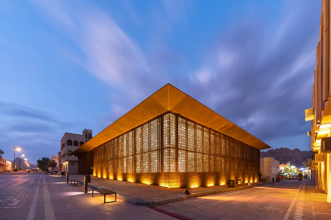 Design Space AlUla al tramonto: facciata in metallo forato illuminata sotto un profondo tetto a sbalzo, vista dalla strada – Design Space AlUla, Arabia Saudita, 2024 – architettura