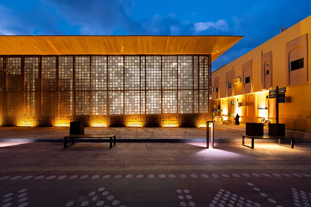 Prospetto principale al tramonto, lo schermo forato illuminato dall'interno accanto a un edificio giallo vicino – Design Space AlUla, Arabia Saudita, 2024 – architettura
