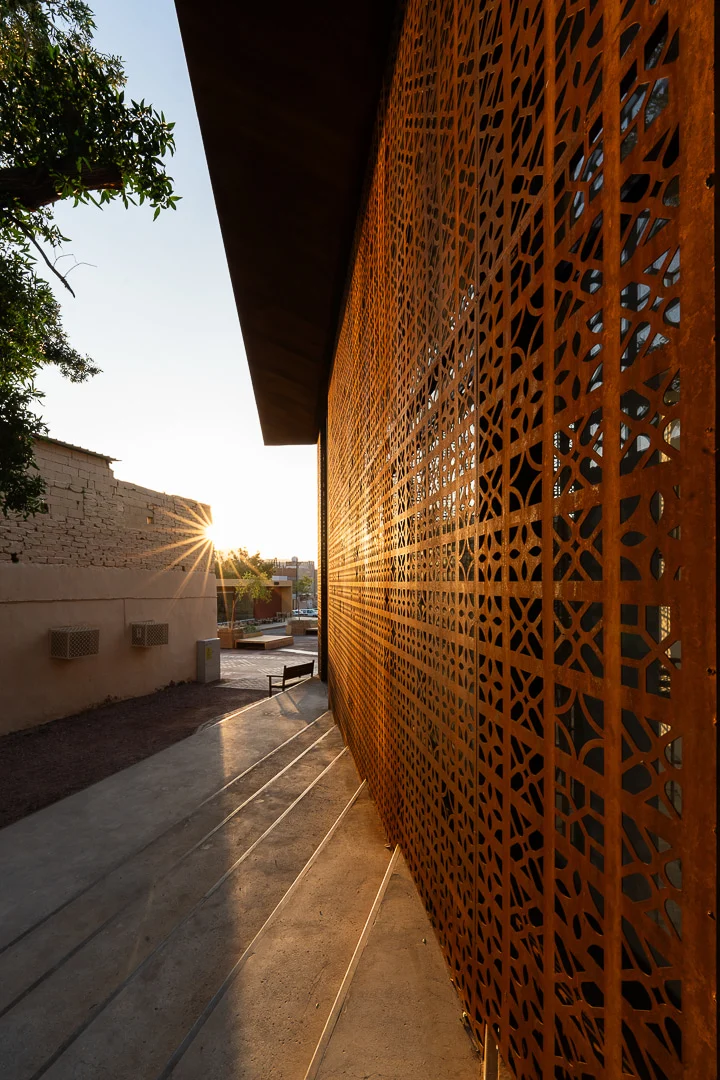 Sole basso che radente illumina lo schermo in metallo forato color ruggine e la pavimentazione in cemento a gradoni – Design Space AlUla, Arabia Saudita, 2024 – architettura
