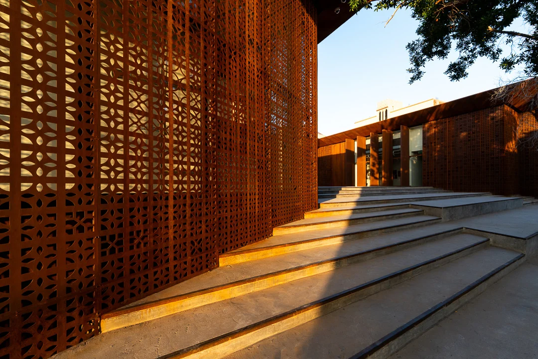 Ampi gradini in cemento lungo lo schermo forato illuminato dal sole, che portano all'ingresso coperto – Design Space AlUla, Arabia Saudita, 2024 – architettura