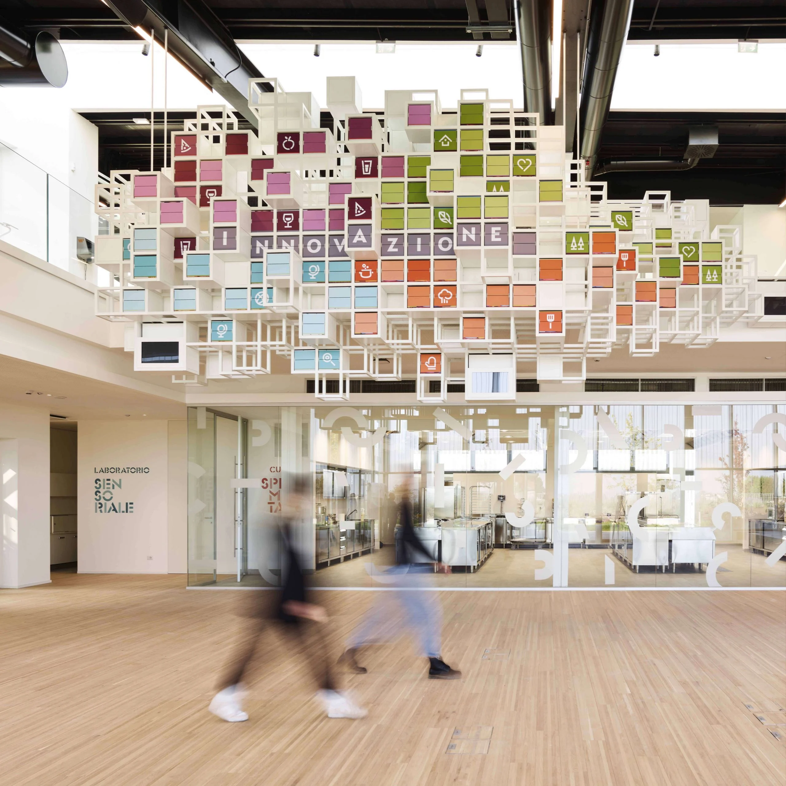 Visitatori sotto La Nuvola, una nuvola sospesa di cubi colorati a palette che compongono la parola "Innovazione" – La Nuvola al Cirfood District, Reggio Emilia, Italia, 2022 – progettazione di installazioni cinetiche
