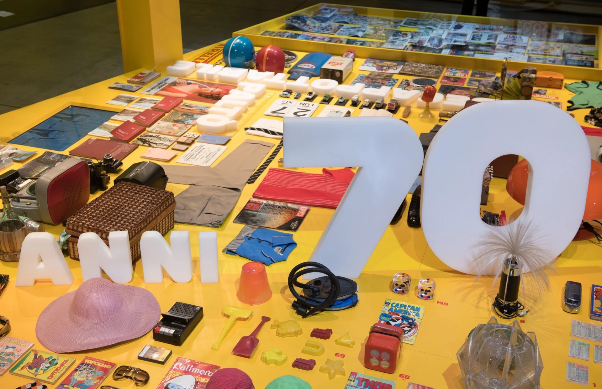 Dettaglio del tavolo espositivo giallo con la scritta Anni 70 tra cappelli, giocattoli, vestiti e oggetti degli anni Settanta – Esselunga Supermostra 60° anniversario, Milano, Italia, 2018 – allestimento espositivo