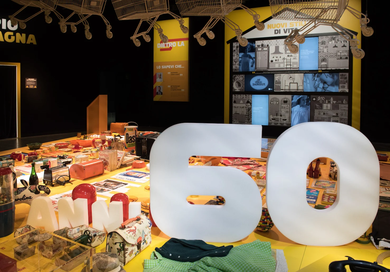 Grande scritta 60 bianca su un tavolo giallo coperto di oggetti quotidiani, con carrelli appesi sopra – Esselunga Supermostra 60° anniversario, Milano, Italia, 2018 – allestimento espositivo