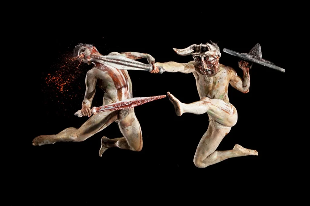 Due guerrieri dipinti d'argilla con spada e scudo si scontrano in aria, uno con un elmo cornuto – I Shardana al Teatro Lirico di Cagliari, Italia, 2013 – scenografia d'opera