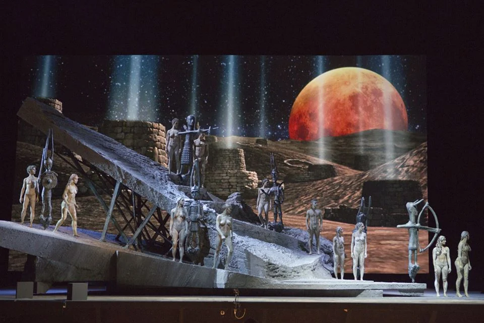 Artisti coperti d'argilla su una scenografia di roccia inclinata davanti a un paesaggio desertico proiettato e a un'enorme luna rossa – I Shardana al Teatro Lirico di Cagliari, Italia, 2013 – scenografia d'opera
