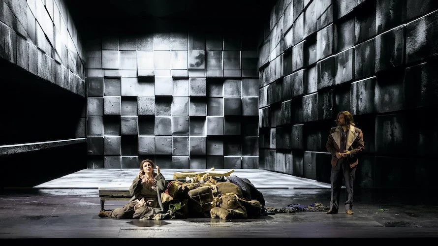 Soprano seduta accanto a una pila di sacchi in una spoglia stanza di blocchi di pietra grigia, un cantante che osserva da destra – Il Trovatore al Teatro Comunale Nouveau, Bologna, Italia, 2024 – scenografia d'opera