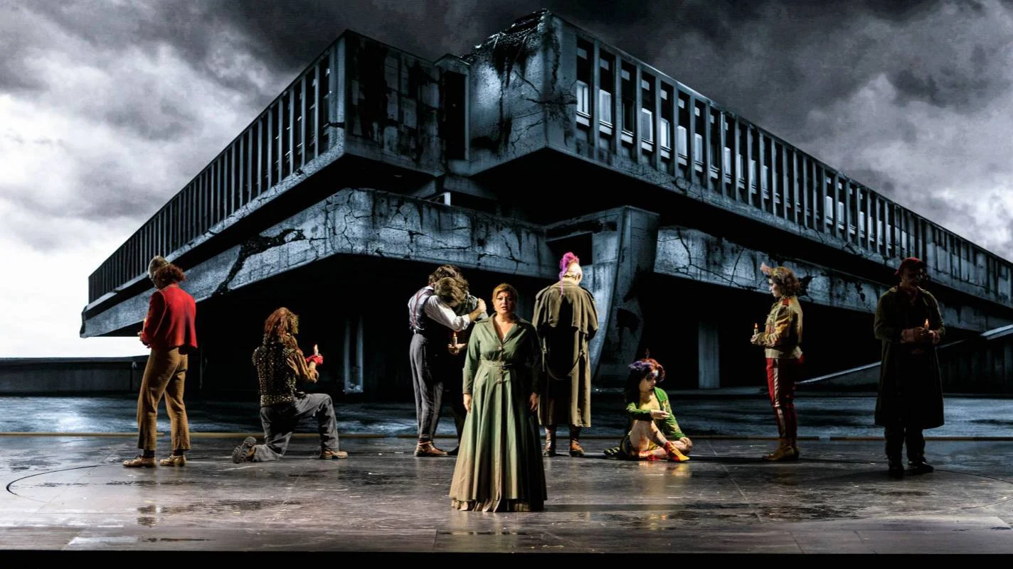 Cantanti in piedi su un palco bagnato davanti a un enorme edificio brutalista in cemento sotto un cielo in tempesta – Il Trovatore al Teatro Comunale Nouveau, Bologna, Italia, 2024 – scenografia d'opera