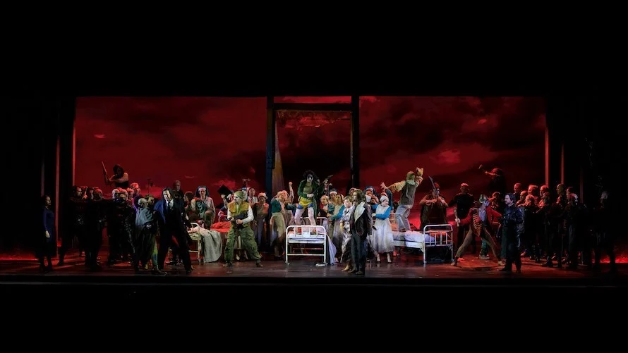 Coro che affolla una scenografia con letti d'ospedale davanti a un fondale di cielo rosso sangue – Il Trovatore al Teatro Comunale Nouveau, Bologna, Italia, 2024 – scenografia d'opera