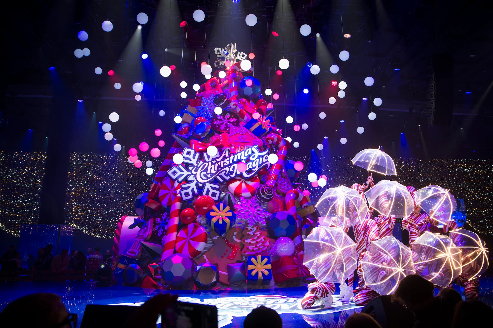 Ballerini con ombrelli luminosi accanto al gigantesco albero di Natale sotto sfere di luce bianca sospese – A Christmas Magic all'Allianz MiCo, Milano, Italia, 2023 – progettazione di eventi
