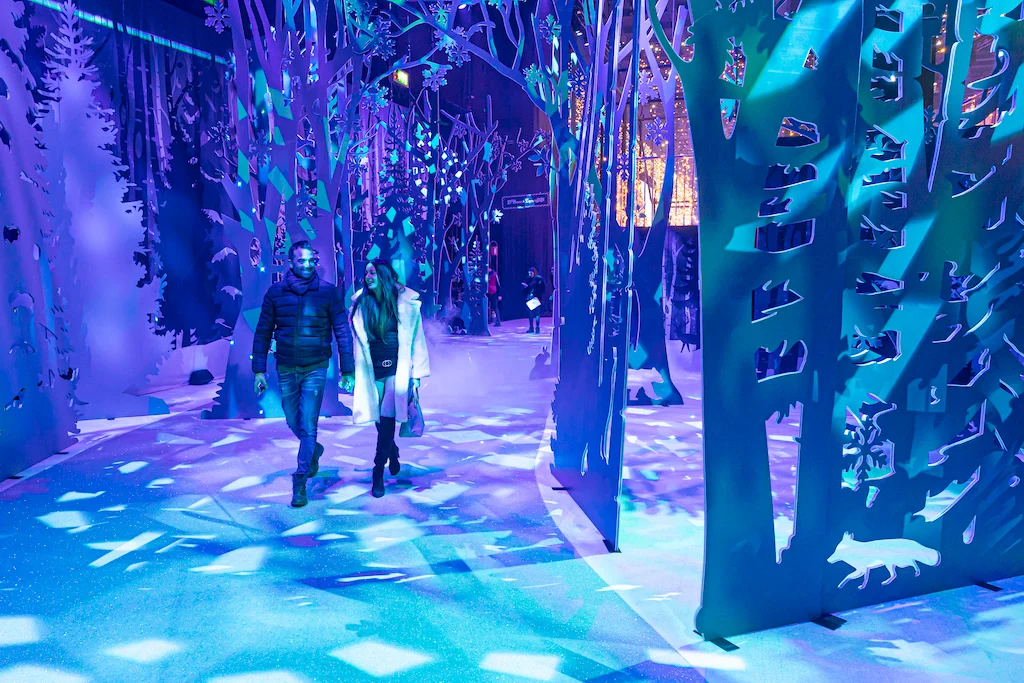 Visitatori che attraversano una foresta di pannelli sagomati ad albero illuminati di blu con proiezioni di fiocchi di neve – A Christmas Magic all'Allianz MiCo, Milano, Italia, 2023 – progettazione di eventi