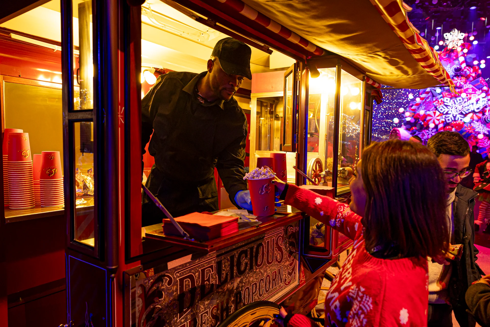 Venditore che serve popcorn da un carretto rosso d'epoca a visitatori con maglioni natalizi – A Christmas Magic all'Allianz MiCo, Milano, Italia, 2023 – progettazione di eventi