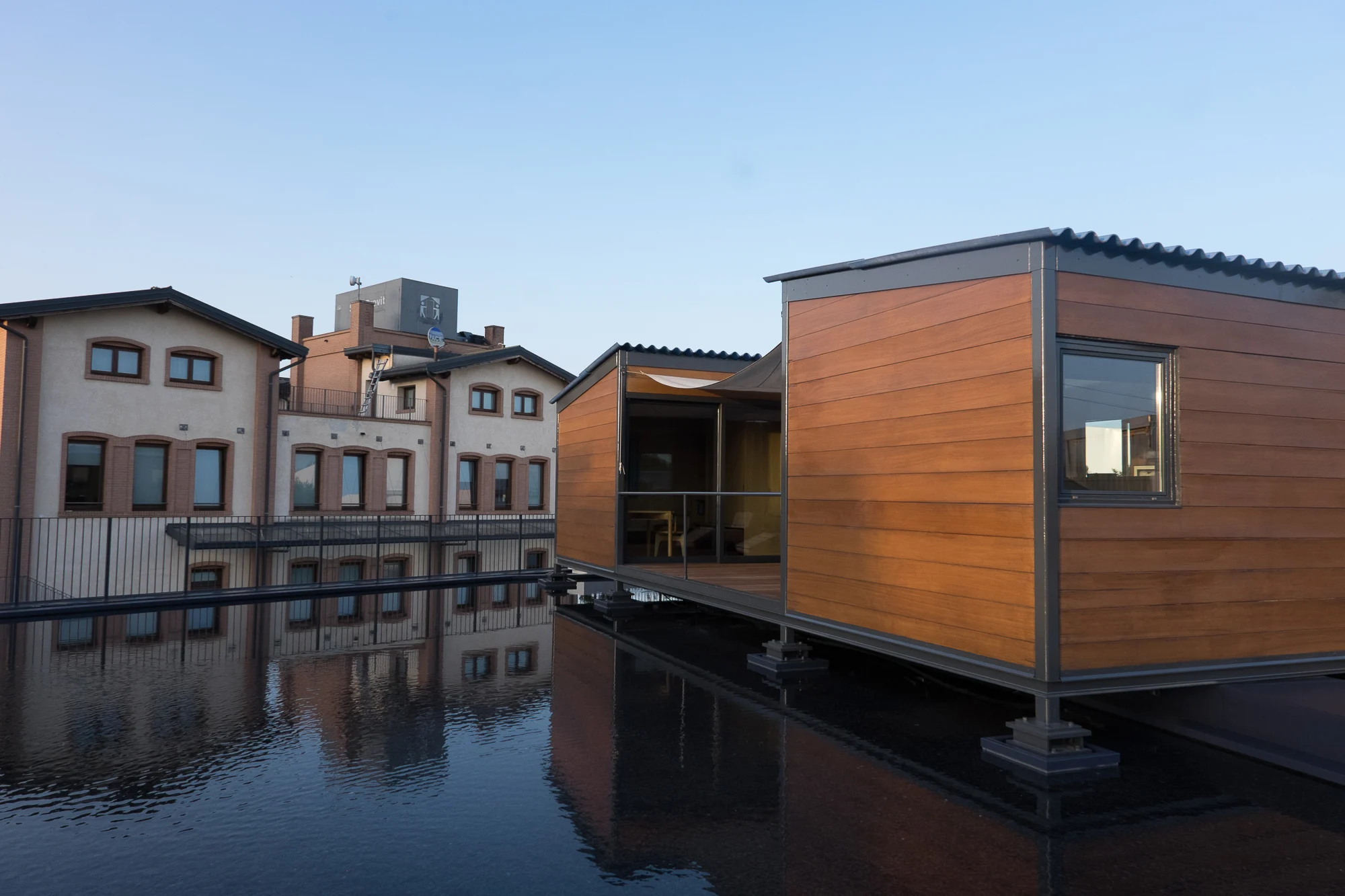 Padiglione rivestito in legno con tetto in lamiera ondulata su palafitte sopra una vasca a specchio, accanto agli edifici in mattoni della fabbrica – Factory Reflections presso Sice Previt, Segrate, Italia, 2024 – allestimento espositivo