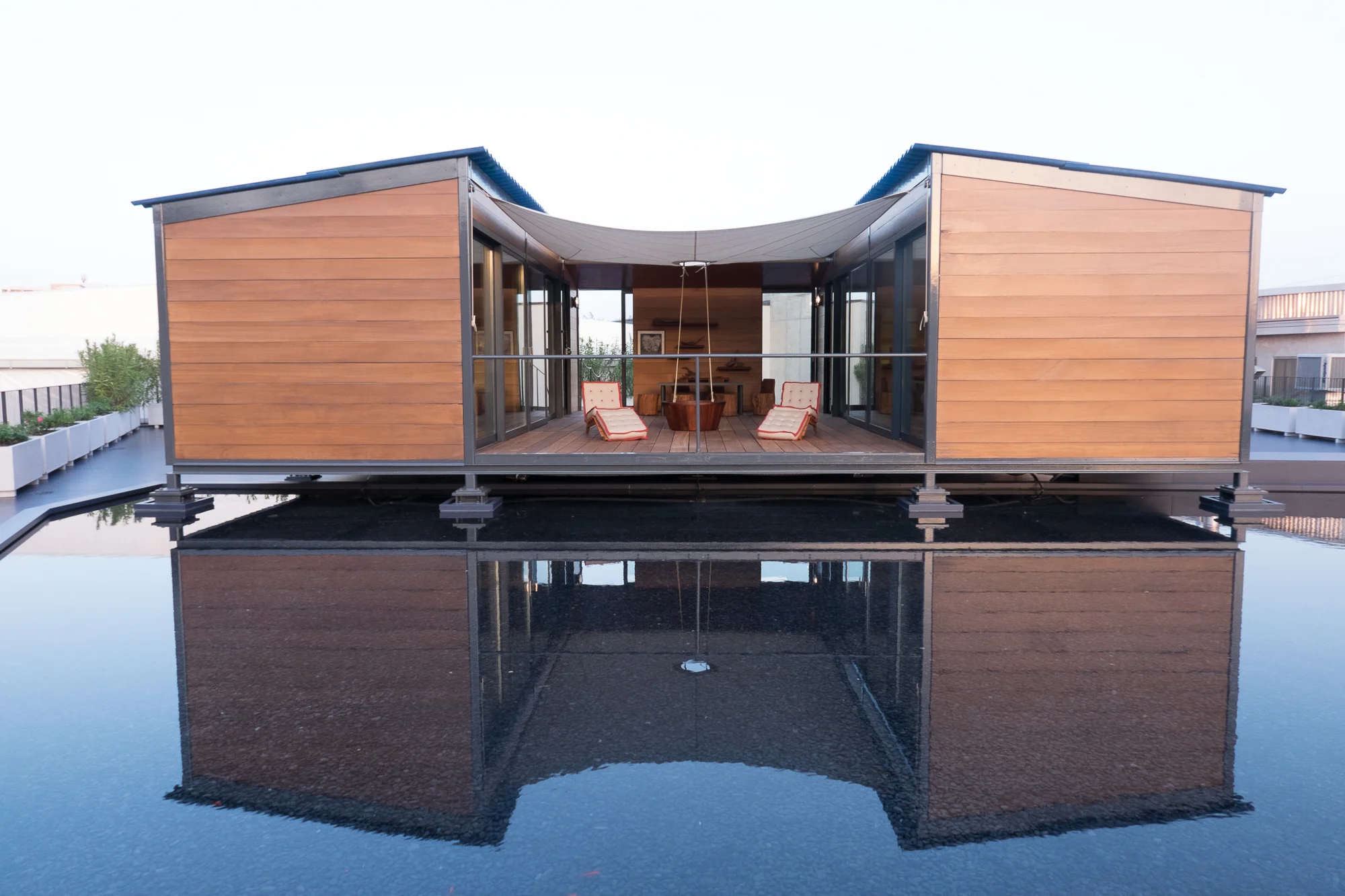 Vista simmetrica del padiglione rivestito in legno con una lounge centrale aperta, riflesso nella vasca d'acqua sul tetto – Factory Reflections presso Sice Previt, Segrate, Italia, 2024 – allestimento espositivo