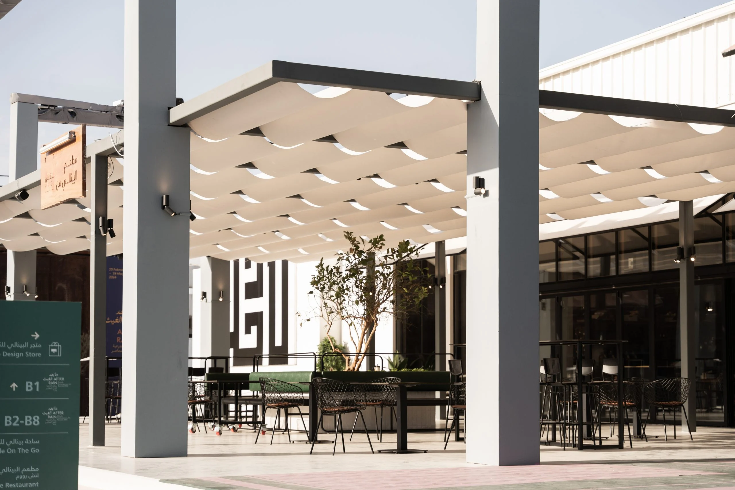 Sedute all'aperto sotto tende in tessuto bianco plissettato su colonne sottili, una grafica in stile cufico dietro – Biennale d'Arte Contemporanea di Diriyah, Riyadh, Arabia Saudita, 2024 – architettura