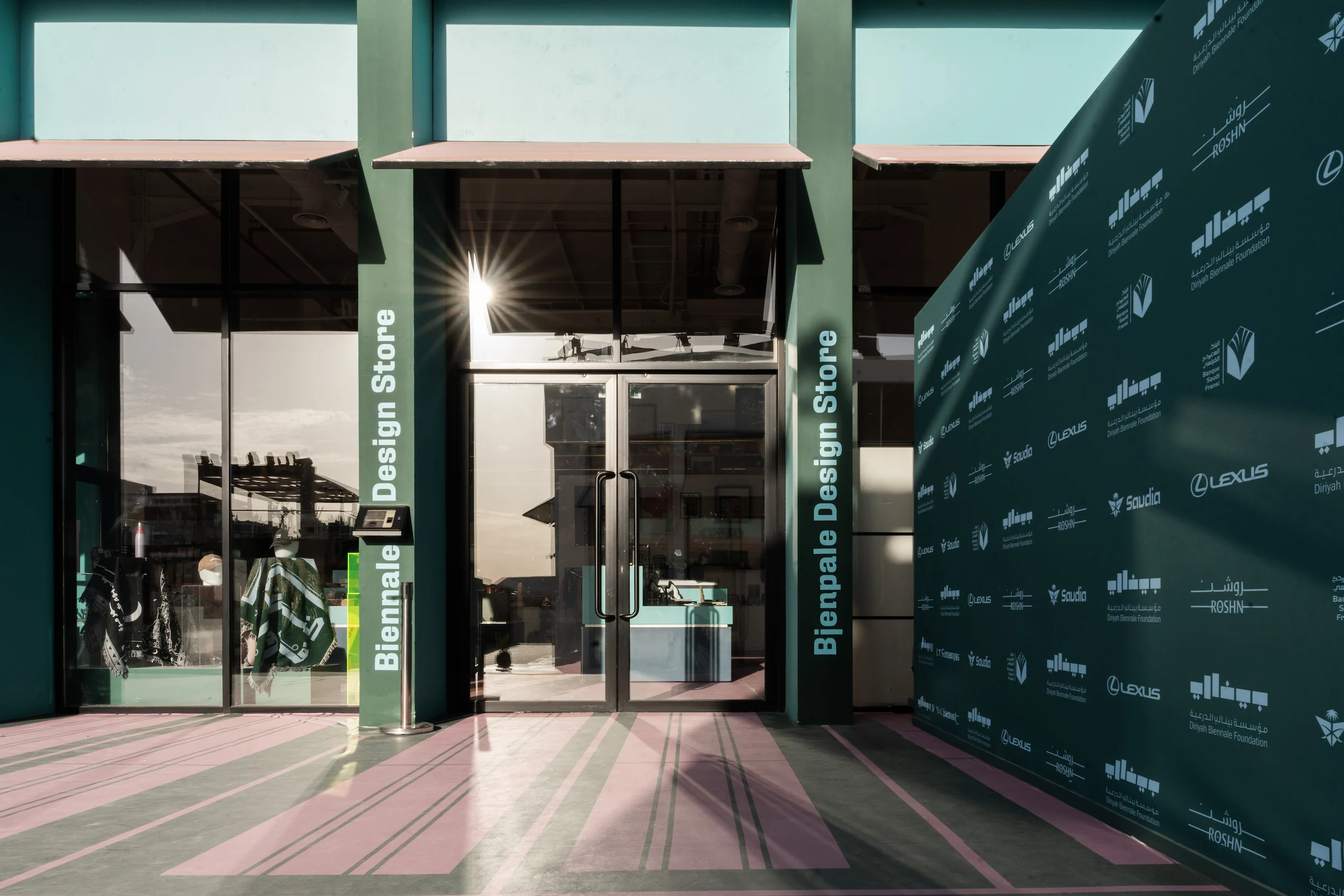 Ingresso verde del Biennale Design Store accanto a una parete degli sponsor, pavimentazione a strisce rosa e verdi – Biennale d'Arte Contemporanea di Diriyah, Riyadh, Arabia Saudita, 2024 – architettura