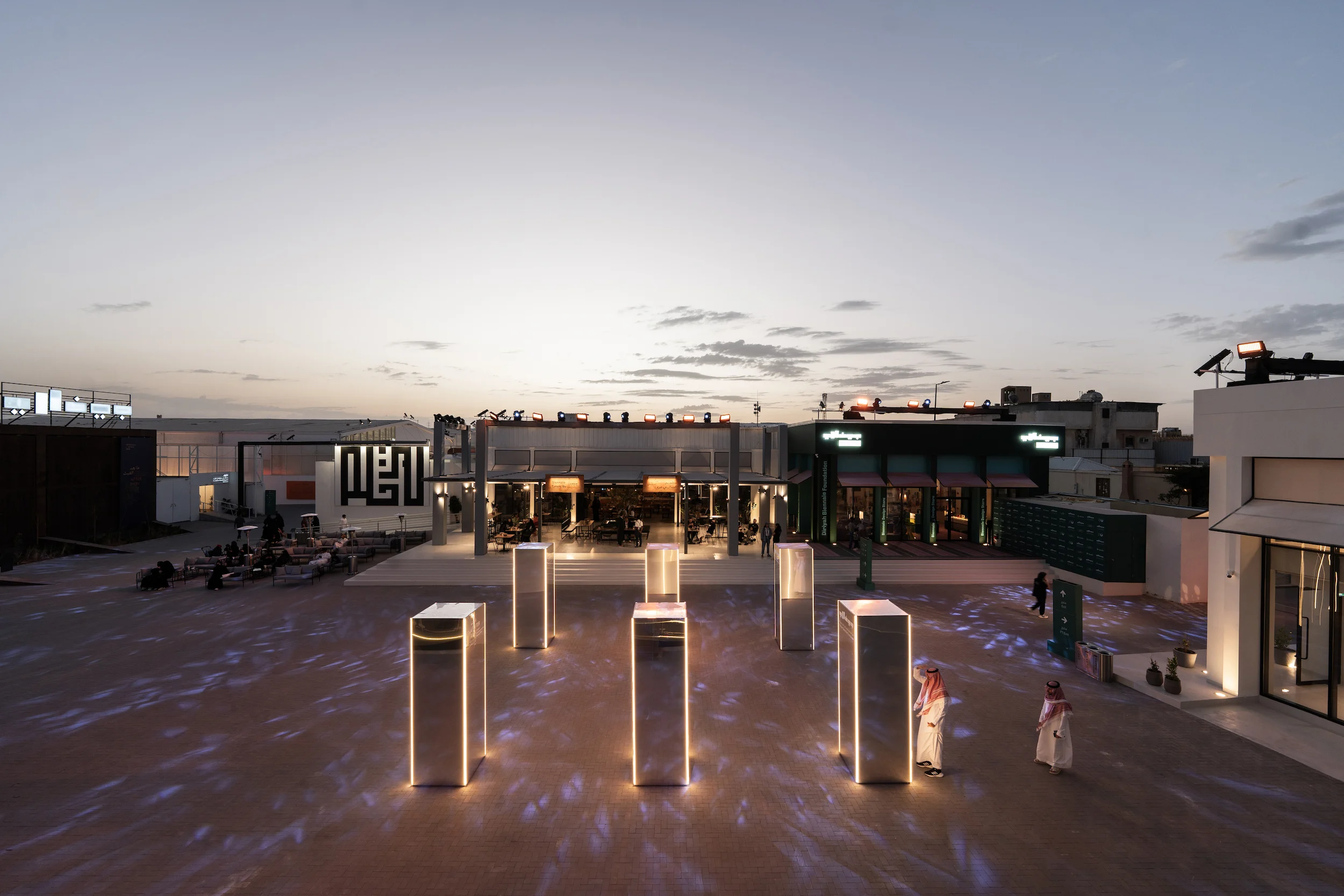 Piazza della Biennale al tramonto con totem a specchio illuminati, nuovi padiglioni e un ristorante sullo sfondo – Biennale d'Arte Contemporanea di Diriyah, Riyadh, Arabia Saudita, 2024 – architettura