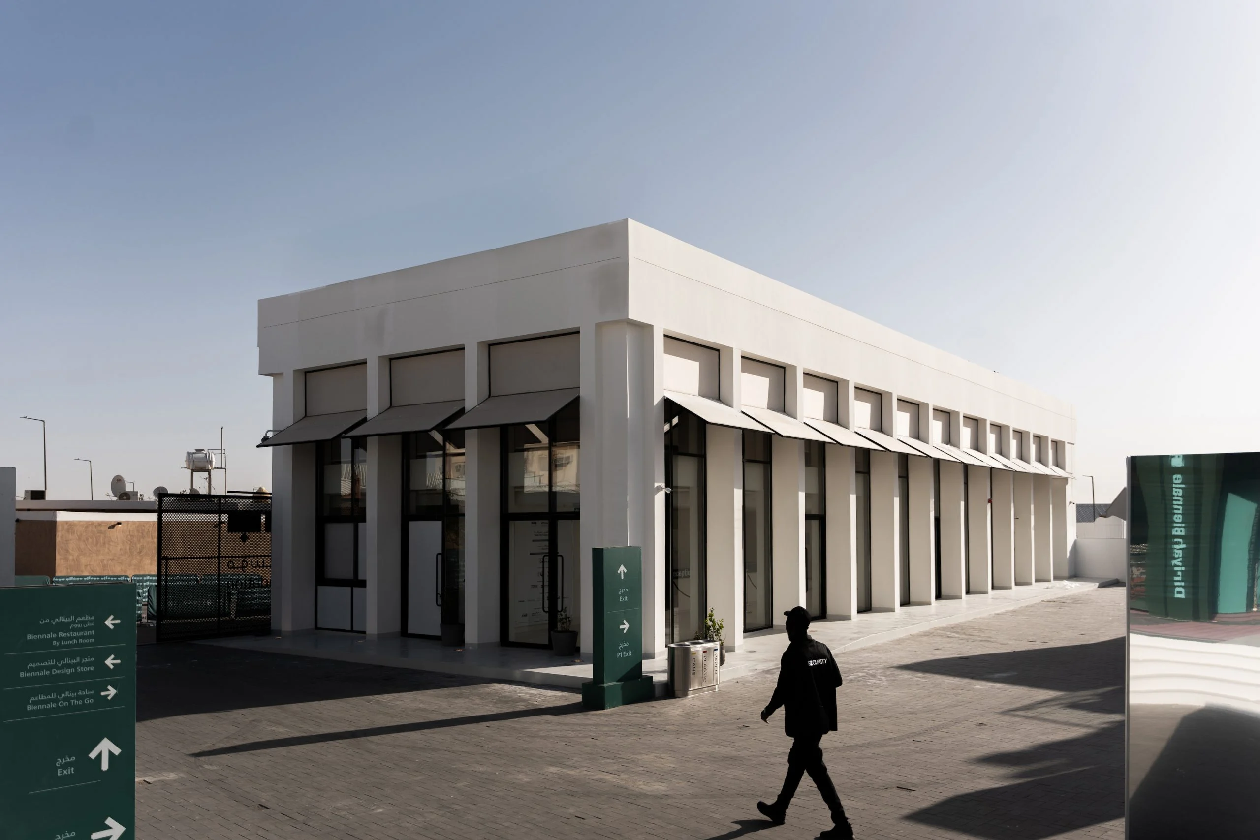 Edificio bianco dell'accoglienza con pilastri sottili e tende, segnaletica verde in primo piano – Biennale d'Arte Contemporanea di Diriyah, Riyadh, Arabia Saudita, 2024 – architettura