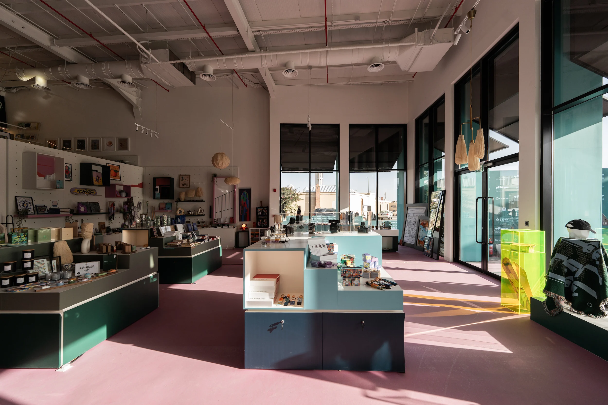 Interno del design store con tavoli espositivi verdi e azzurro chiaro, pavimento rosa e alte finestre – Biennale d'Arte Contemporanea di Diriyah, Riyadh, Arabia Saudita, 2024 – architettura