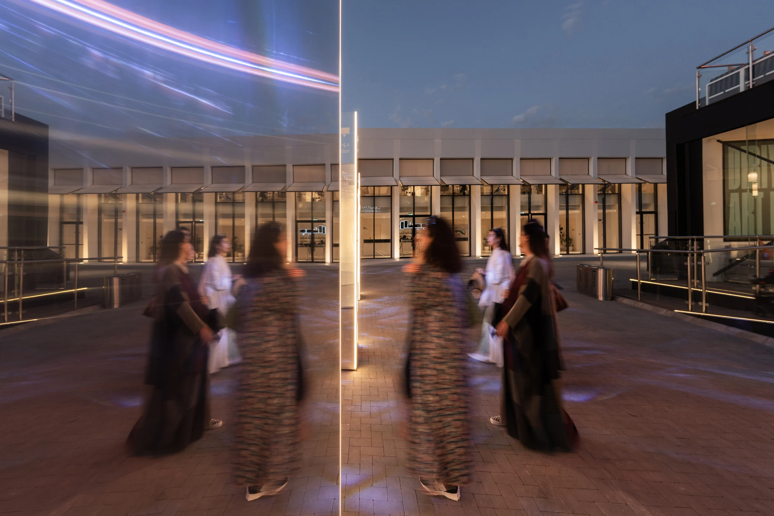 Visitatori riflessi in un totem a specchio nella piazza al tramonto, padiglioni illuminati dietro – Biennale d'Arte Contemporanea di Diriyah, Riyadh, Arabia Saudita, 2024 – architettura