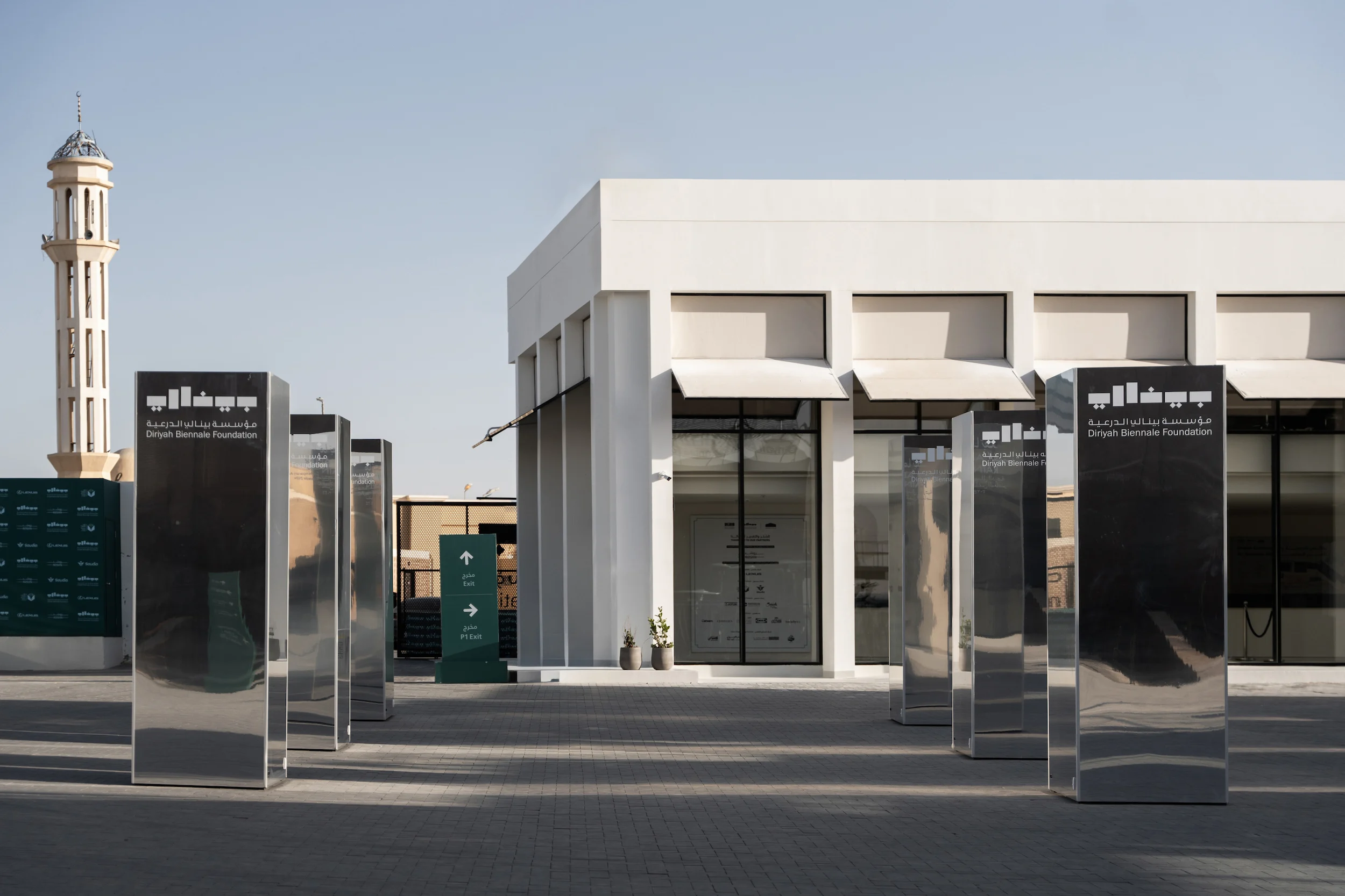 Totem in acciaio a specchio con il logo della Biennale Foundation davanti all'edificio bianco dell'accoglienza, minareto dietro – Biennale d'Arte Contemporanea di Diriyah, Riyadh, Arabia Saudita, 2024 – architettura