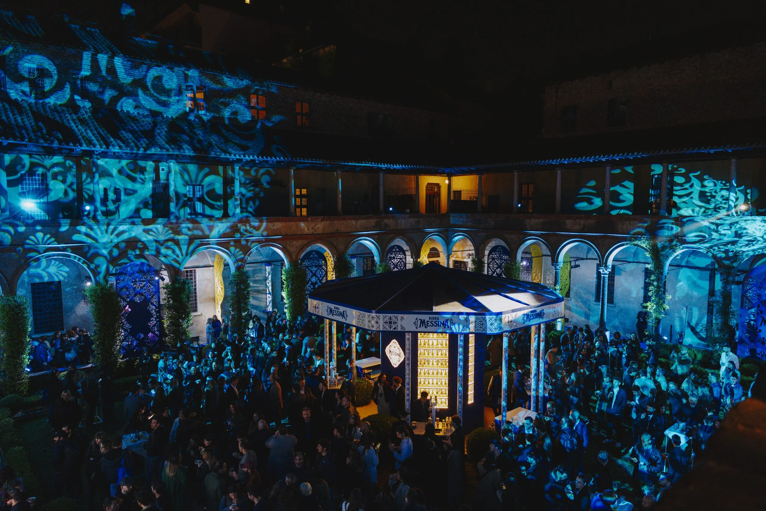 Vista notturna della folla intorno al chiosco, facciate del chiostro coperte da proiezioni blu e turchesi – Birra Messina Bar Meraviglia al Fuorisalone, Milano, Italia, 2024 – progettazione di eventi