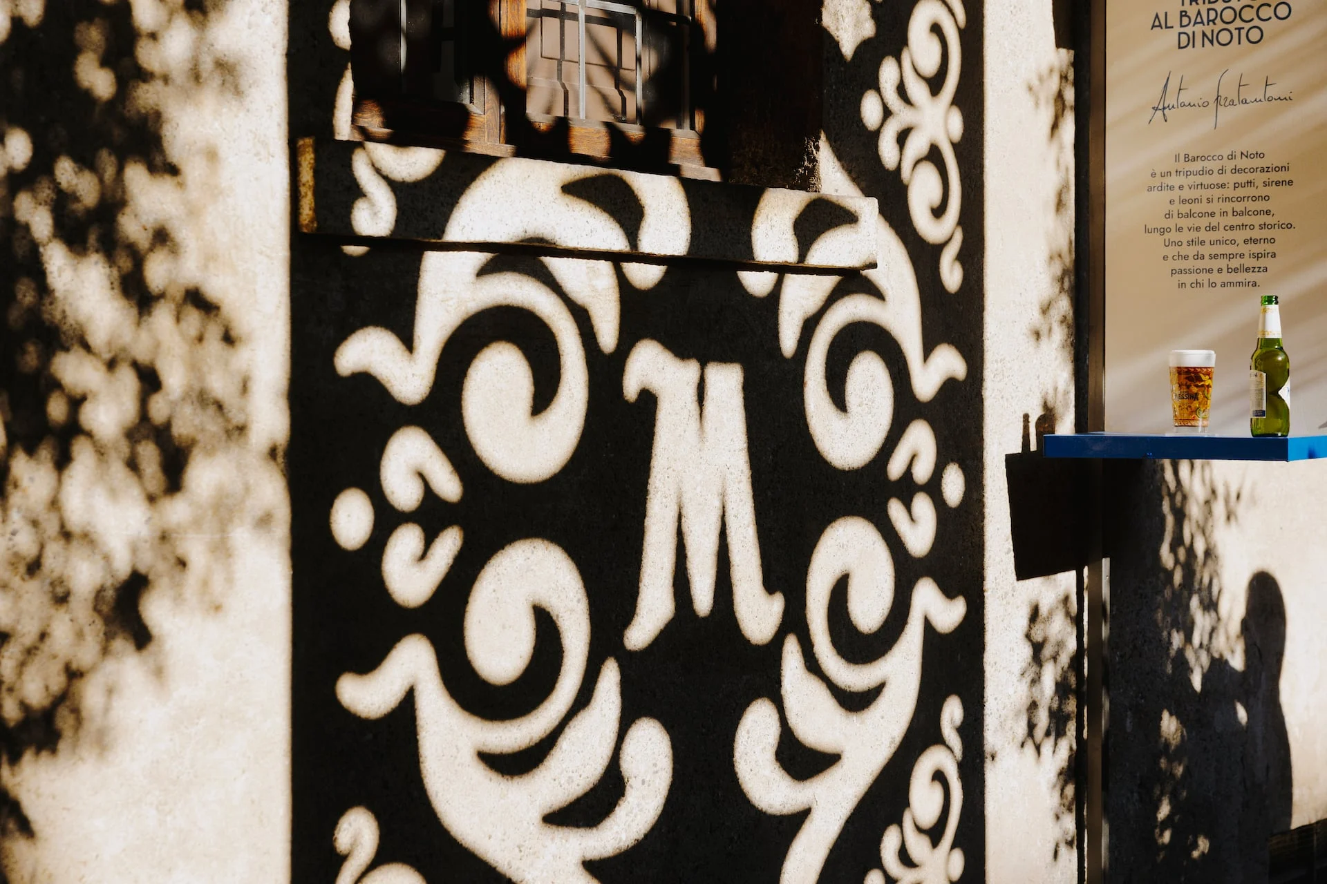 Ombra di un monogramma M traforato proiettata su una parete accanto a una mensola con un bicchiere e una bottiglia di birra – Birra Messina Bar Meraviglia al Fuorisalone, Milano, Italia, 2024 – progettazione di eventi