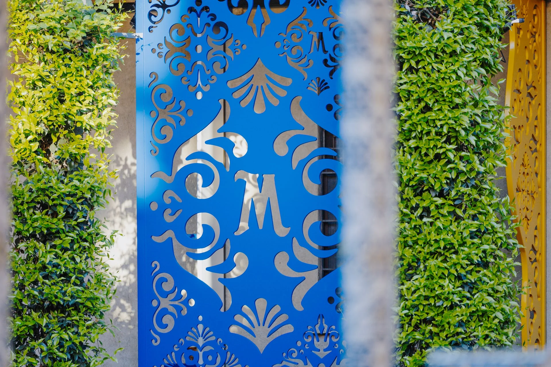 Dettaglio di un pannello metallico traforato blu con monogramma M tra due siepi verdi – Birra Messina Bar Meraviglia al Fuorisalone, Milano, Italia, 2024 – progettazione di eventi