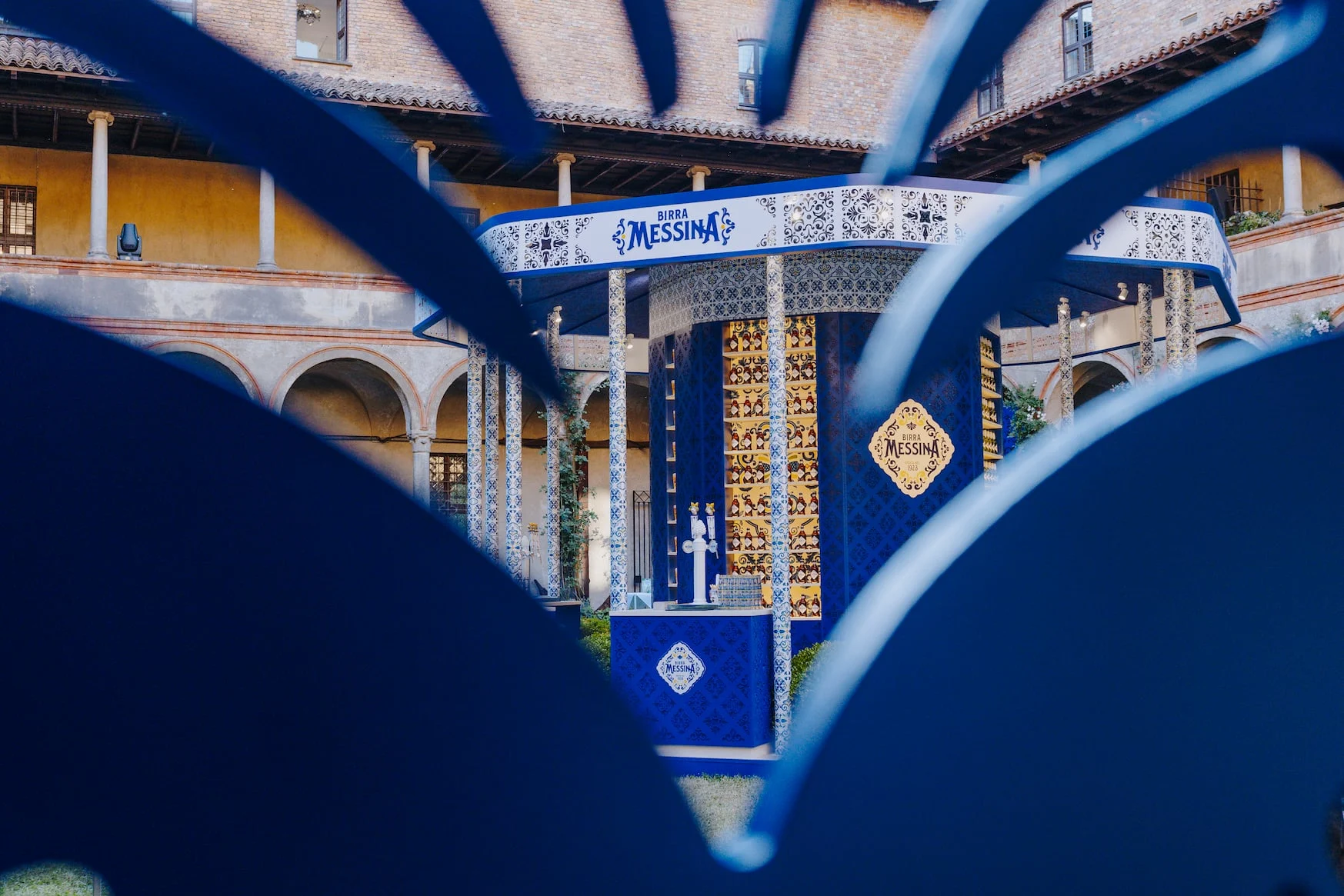 Il chiosco Birra Messina incorniciato da forme ornamentali traforate blu in primo piano – Birra Messina Bar Meraviglia al Fuorisalone, Milano, Italia, 2024 – progettazione di eventi