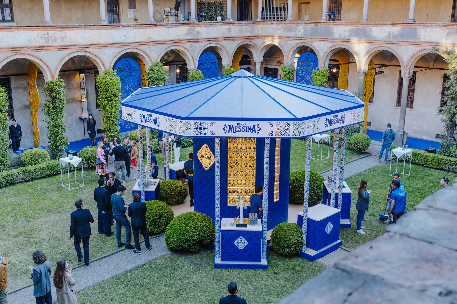 Vista dall'alto di giorno del chiosco blu Birra Messina nel giardino del chiostro, con colonne in stile maiolica – Birra Messina Bar Meraviglia al Fuorisalone, Milano, Italia, 2024 – progettazione di eventi