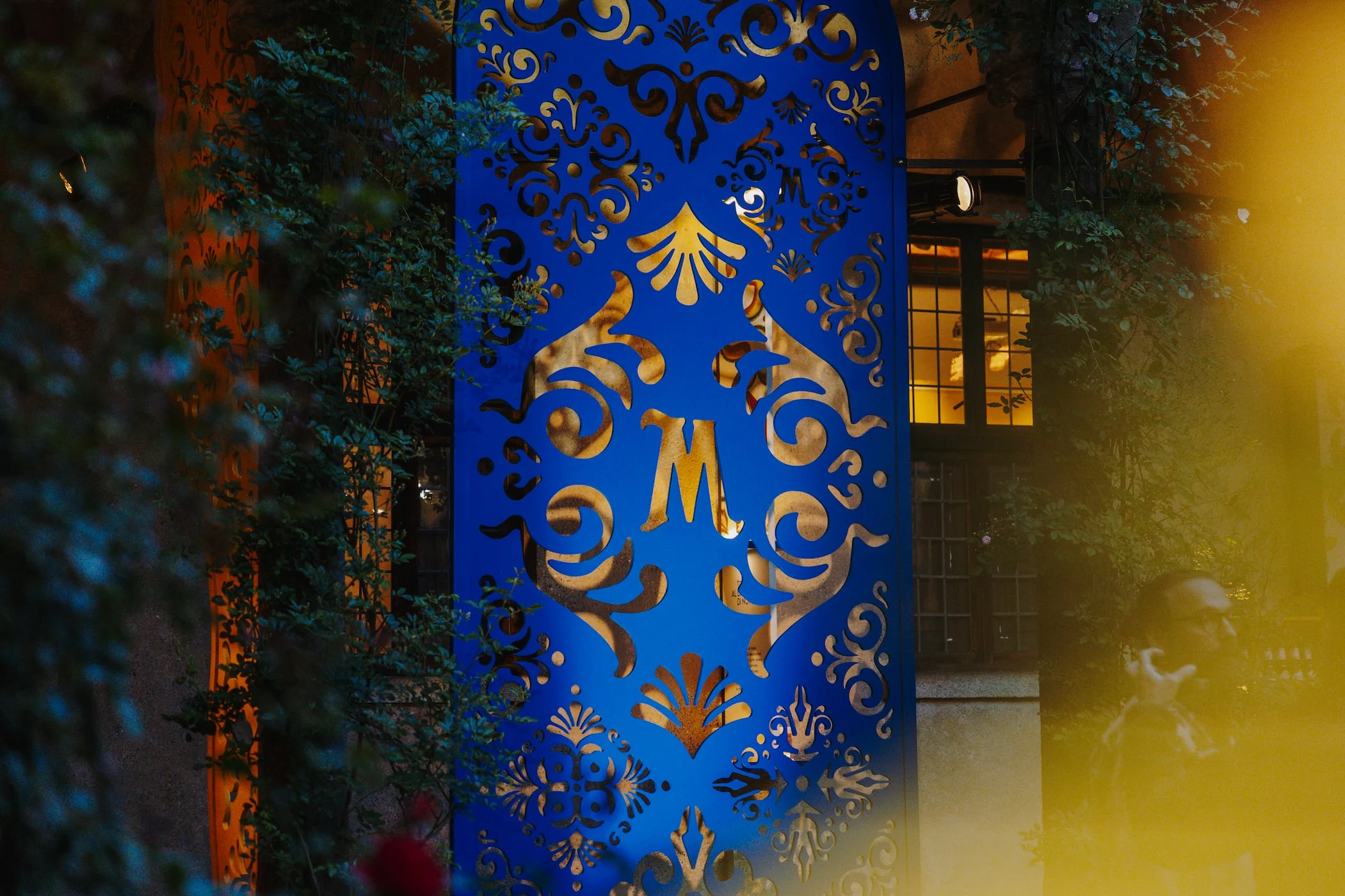 Pannello traforato blu con un monogramma M dorato che brilla di notte tra il fogliame scuro – Birra Messina Bar Meraviglia al Fuorisalone, Milano, Italia, 2024 – progettazione di eventi