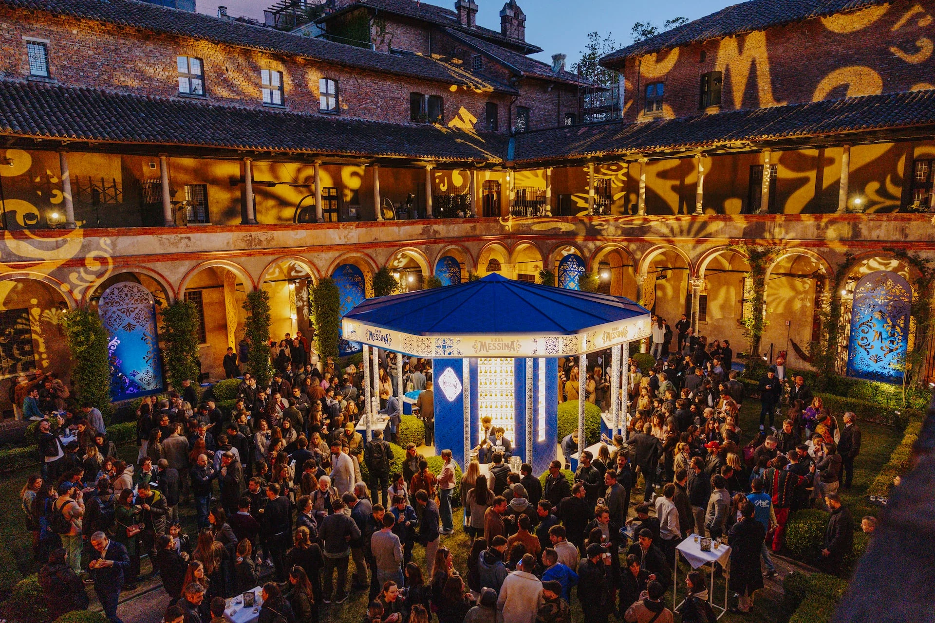Vista notturna dall'alto del chiostro affollato, portici e pareti superiori illuminati da proiezioni di motivi dorati – Birra Messina Bar Meraviglia al Fuorisalone, Milano, Italia, 2024 – progettazione di eventi