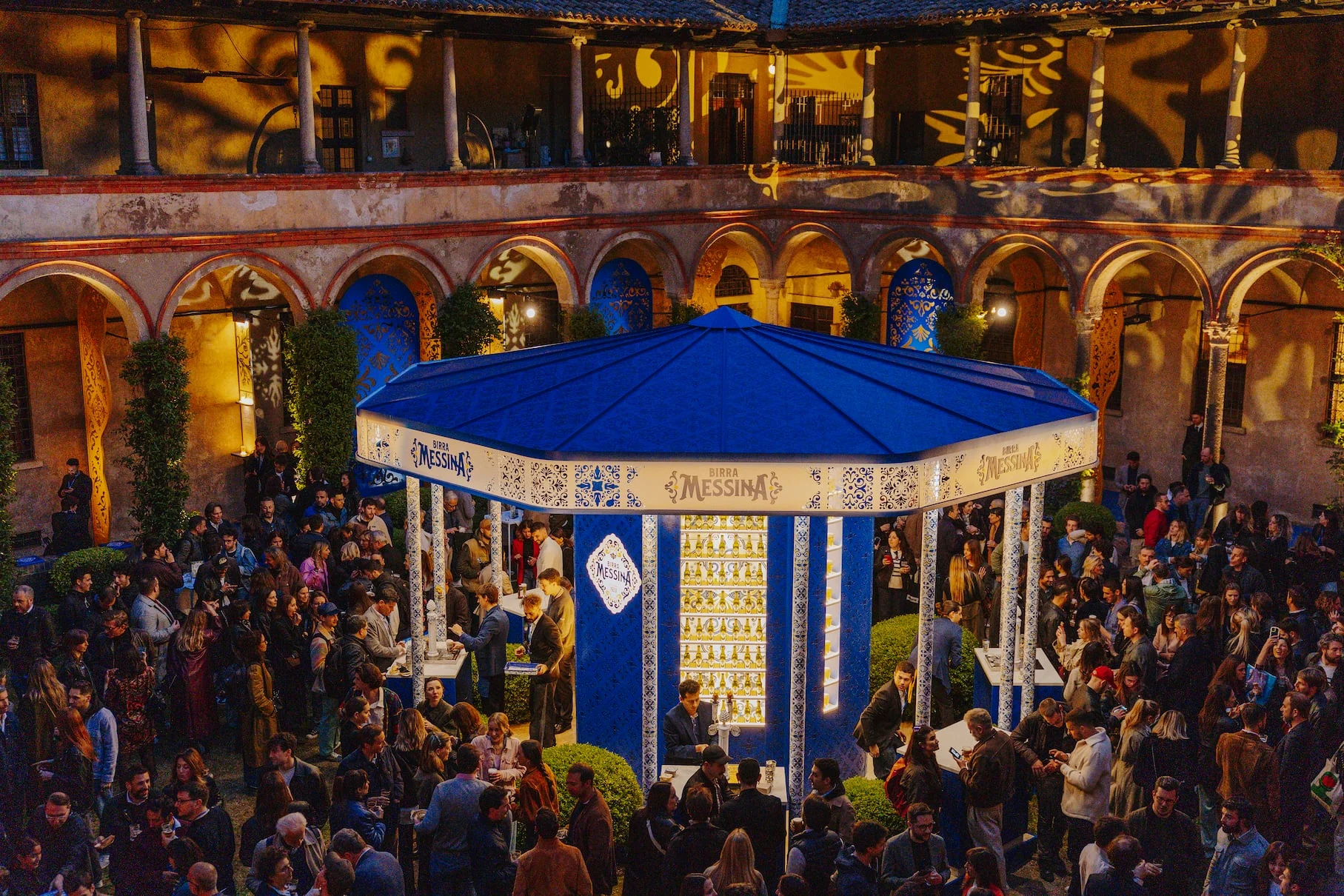 Chiosco bar blu Birra Messina nel chiostro affollato al tramonto, portici avvolti da proiezioni di motivi dorati – Birra Messina Bar Meraviglia al Fuorisalone, Milano, Italia, 2024 – progettazione di eventi