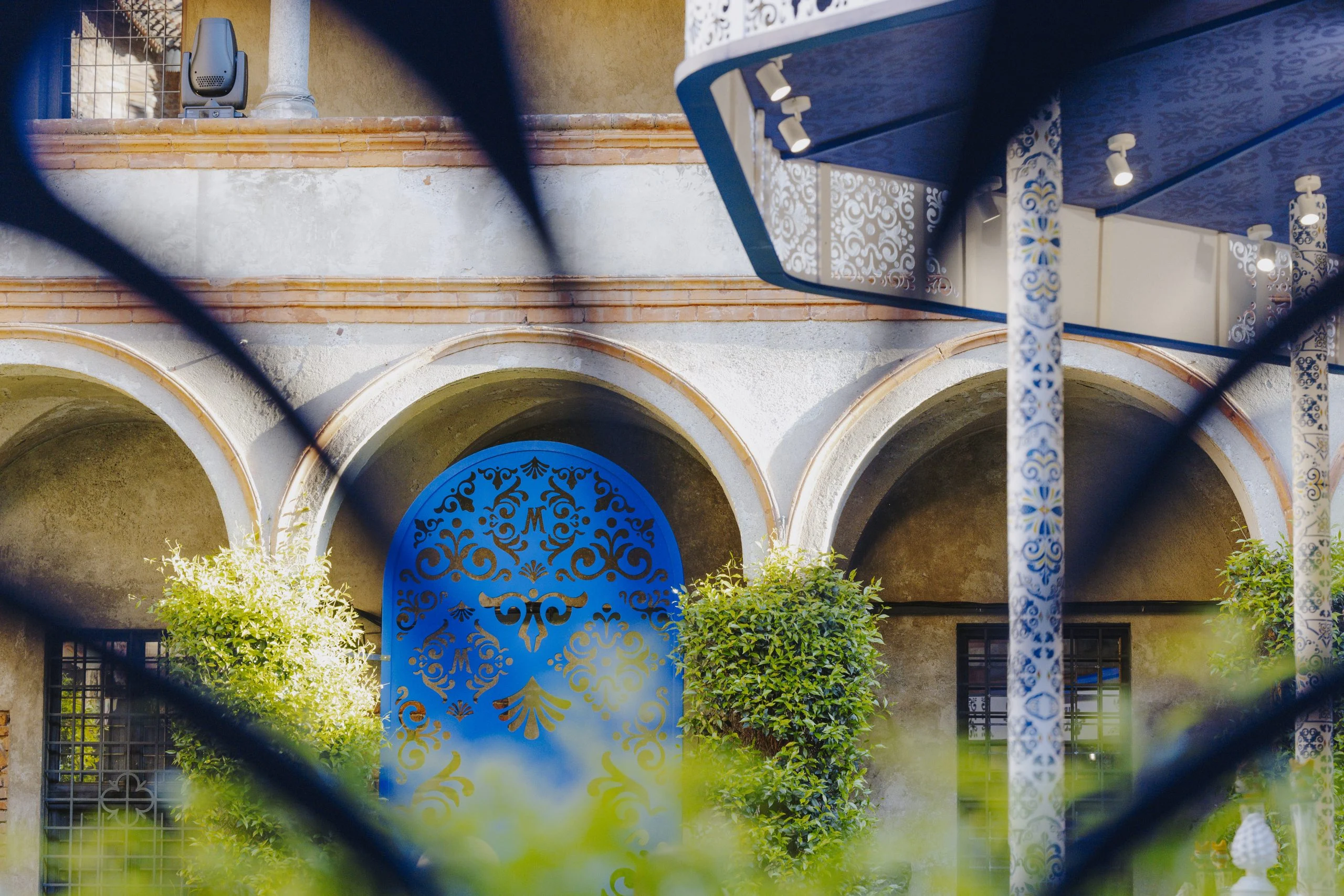 Pannello ornamentale traforato blu in un arco del chiostro, visto attraverso forme blu sfocate in primo piano – Birra Messina Bar Meraviglia al Fuorisalone, Milano, Italia, 2024 – progettazione di eventi