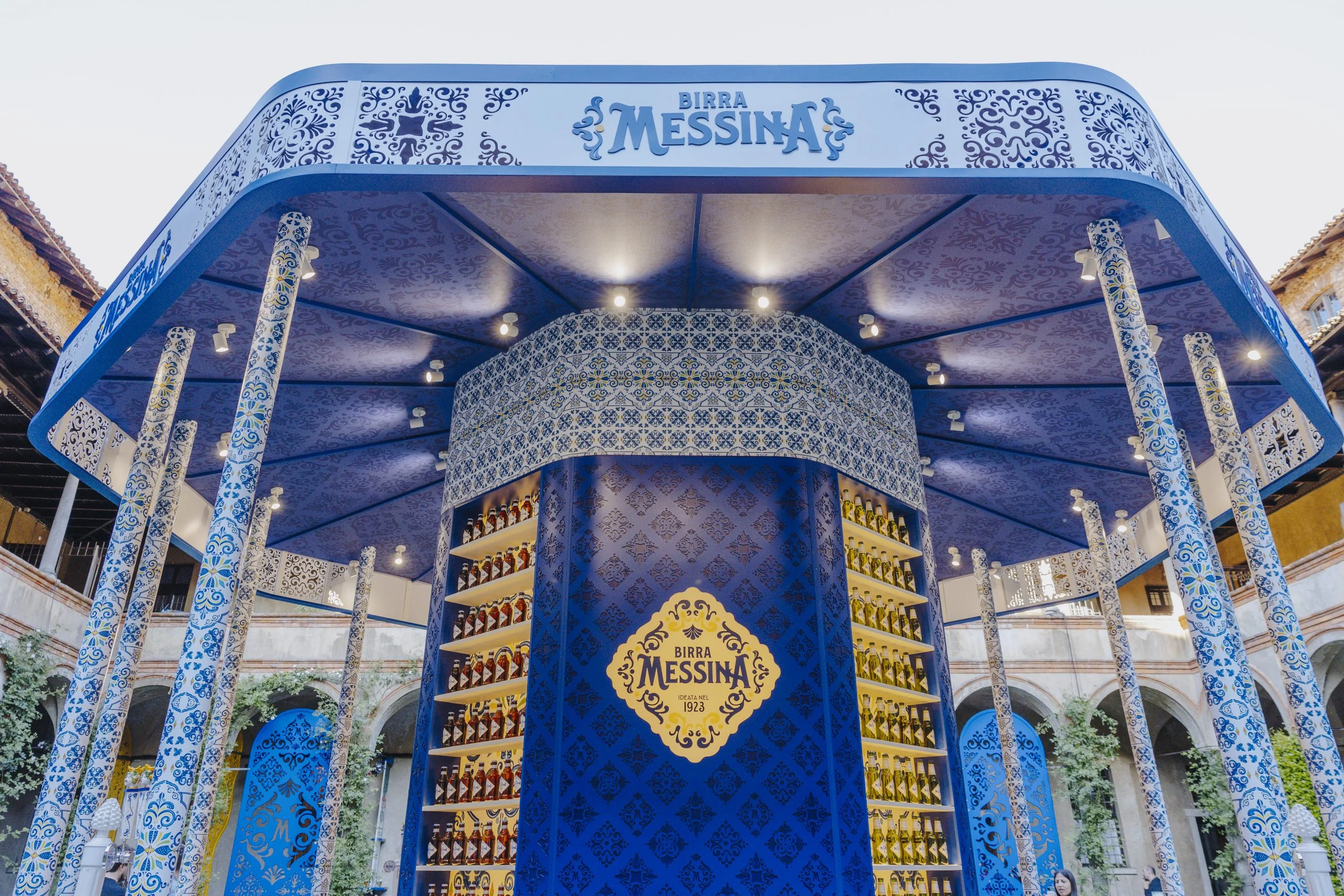 Vista ravvicinata del chiosco Birra Messina: colonne piastrellate, pensilina illuminata e una torre centrale di bottiglie di birra – Birra Messina Bar Meraviglia al Fuorisalone, Milano, Italia, 2024 – progettazione di eventi