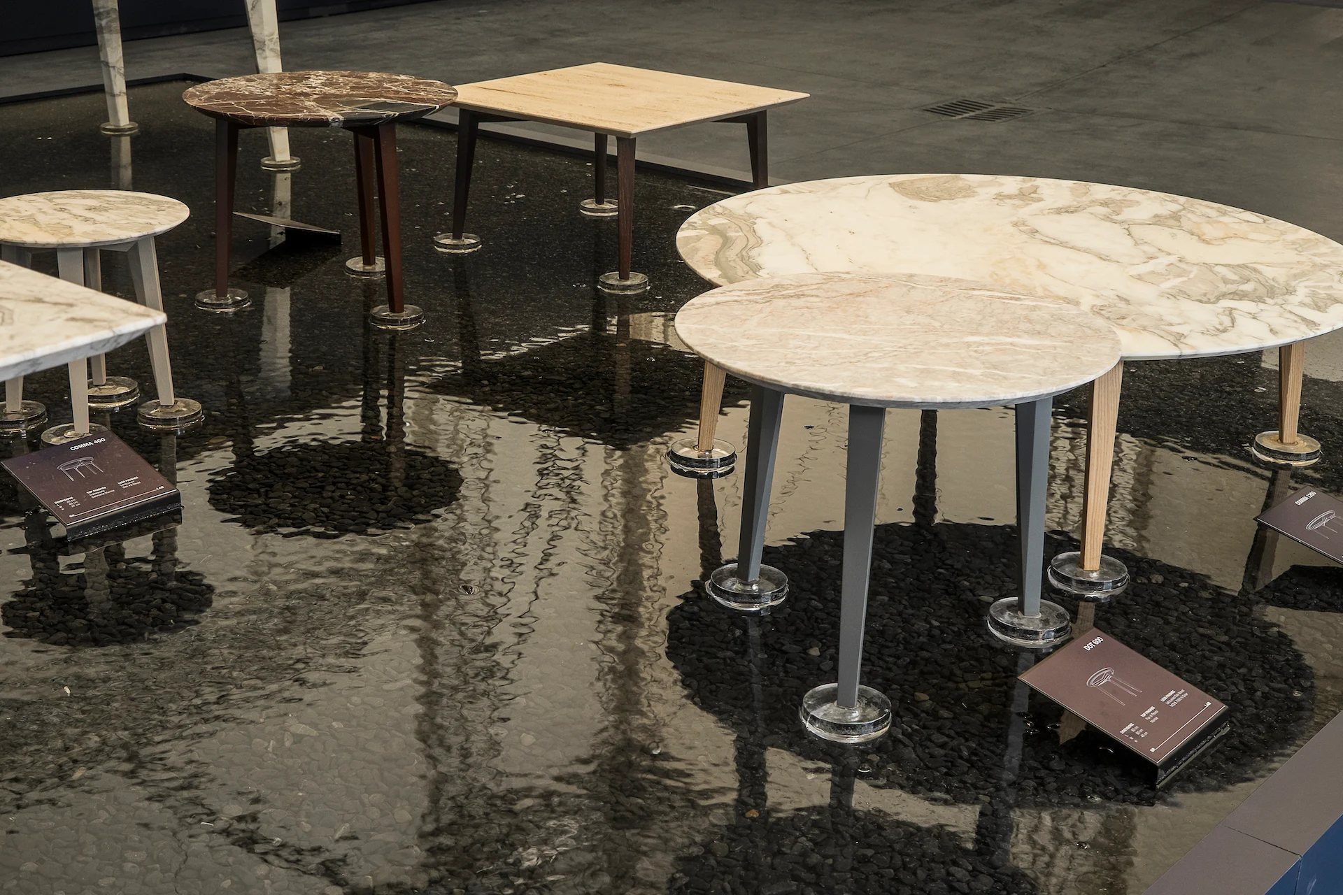 Tavolini rotondi in marmo e legno con piccole targhe informative in un velo d'acqua scura – Factory Reflections presso Sice Previt, Segrate, Italia, 2024 – allestimento espositivo