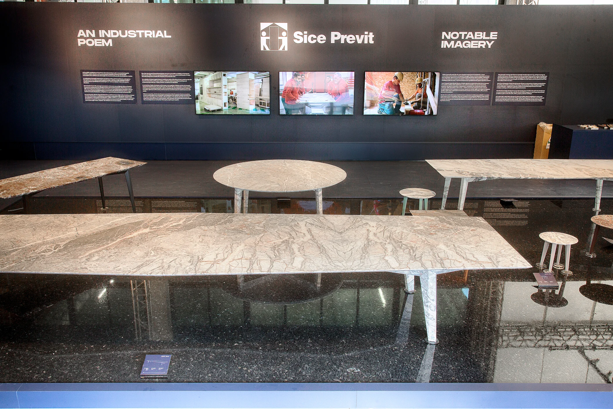 Vista frontale della mostra: tavoli in marmo in una vasca nera di fronte alla parete Sice Previt con schermi e testi – Factory Reflections presso Sice Previt, Segrate, Italia, 2024 – allestimento espositivo