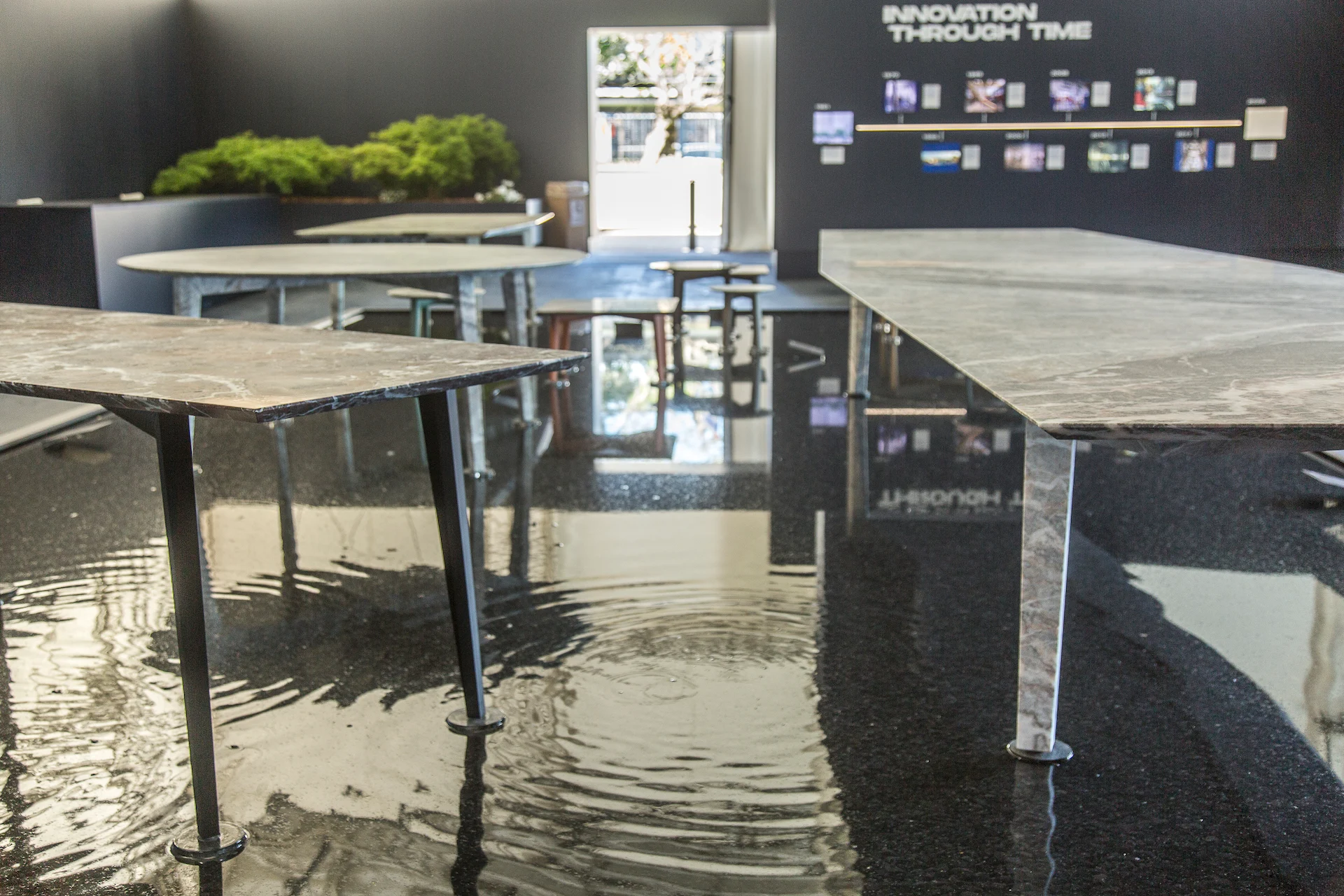 Tavoli in marmo che emergono dall'acqua increspata, con una fioriera di muschio e la parete Innovation Through Time dietro – Factory Reflections presso Sice Previt, Segrate, Italia, 2024 – allestimento espositivo