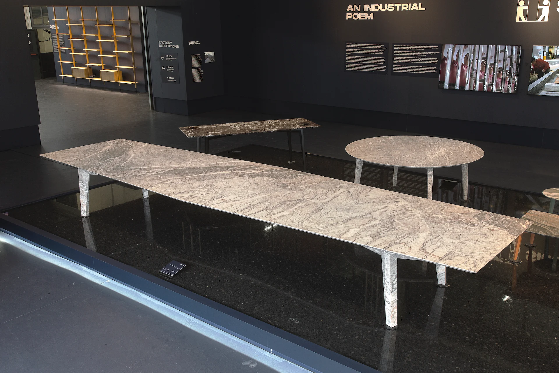 Lungo tavolo in marmo con gambe in acciaio rastremate su un pavimento nero riflettente, sotto la scritta a parete An Industrial Poem – Factory Reflections presso Sice Previt, Segrate, Italia, 2024 – allestimento espositivo