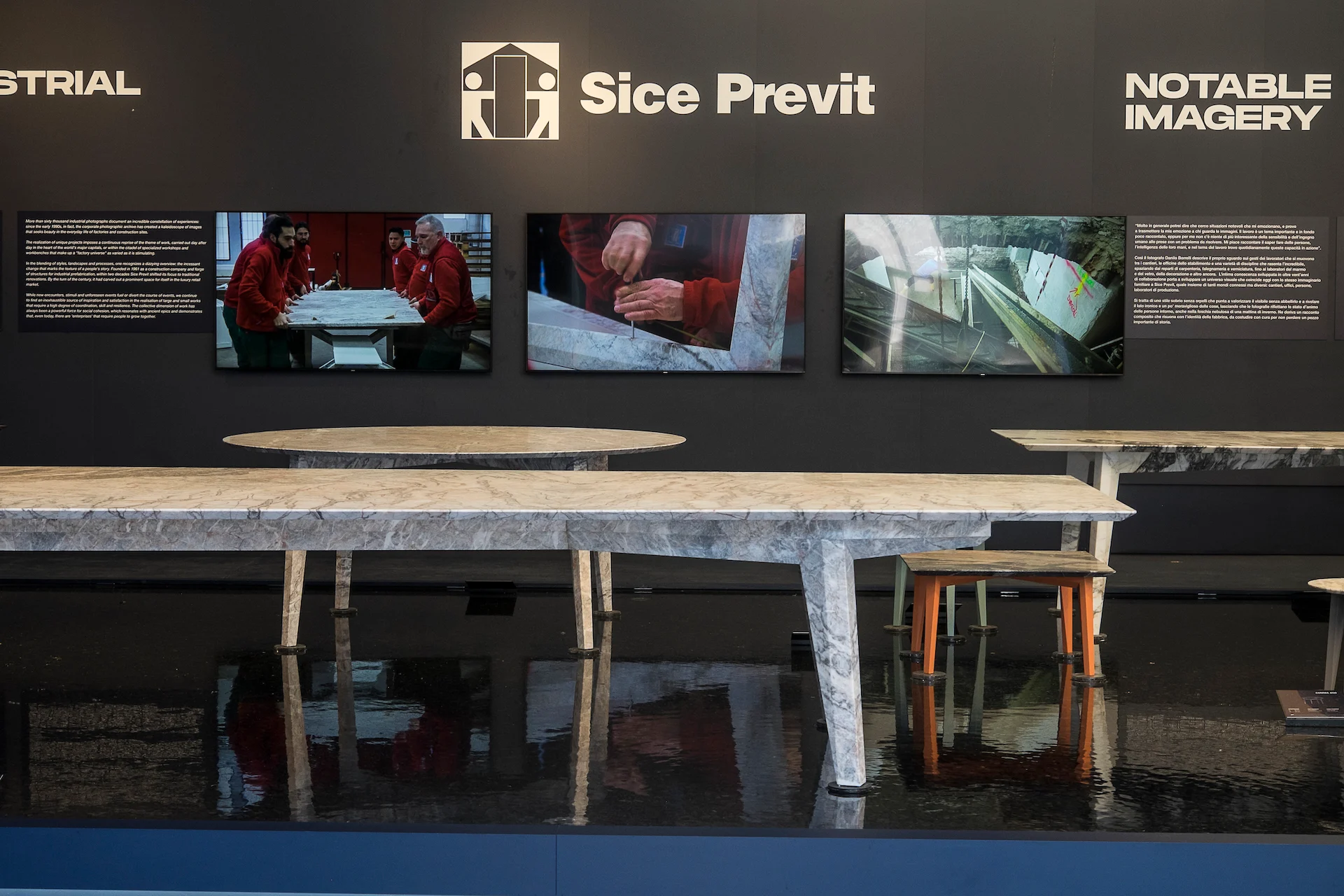 Parete espositiva Sice Previt con schermi video sopra tavoli dal piano in marmo in una vasca nera riflettente – Factory Reflections presso Sice Previt, Segrate, Italia, 2024 – allestimento espositivo